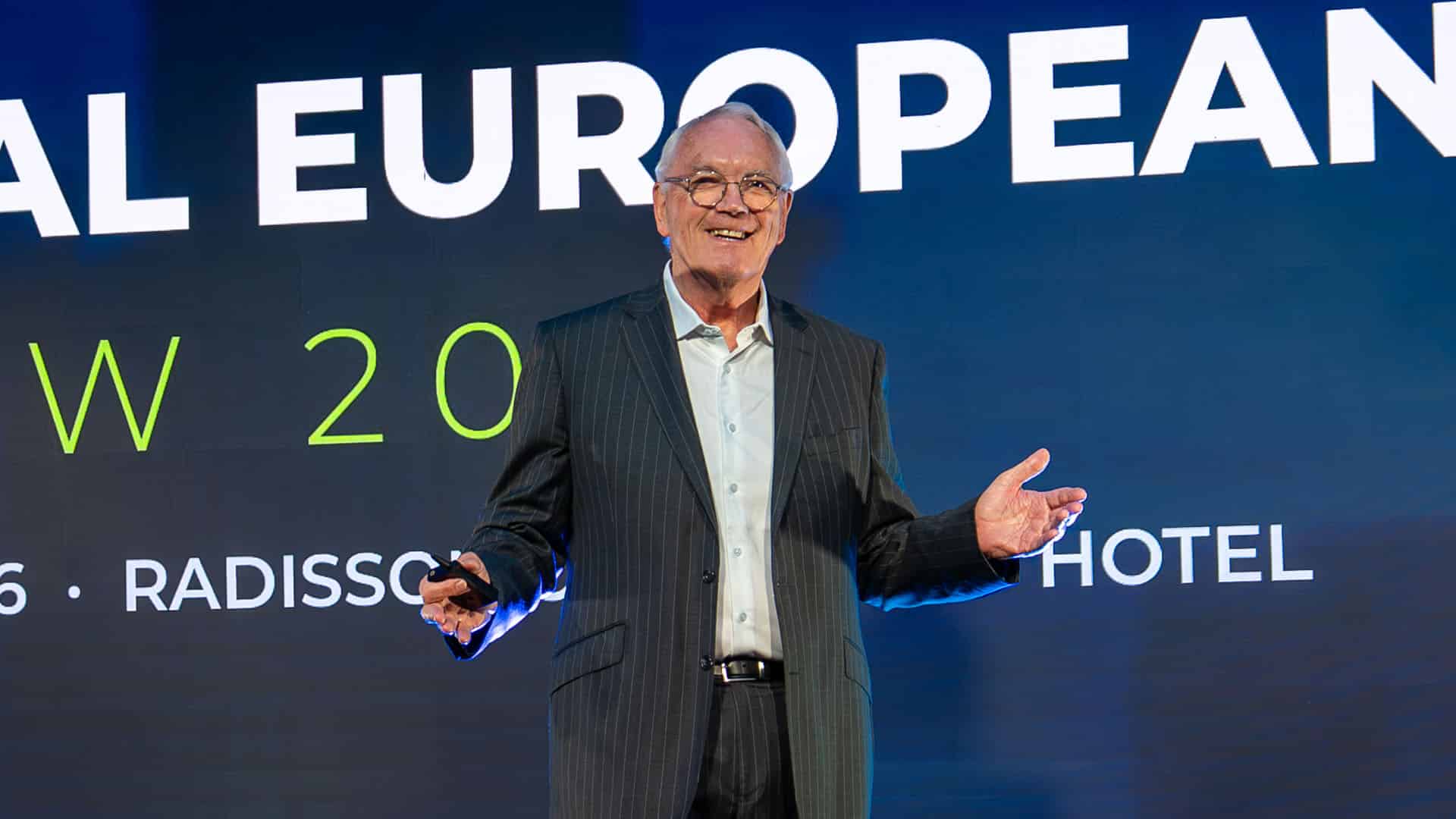 October Advertising Smiling male speaker in a pinstripe suit presenting on a stage with large white text behind him