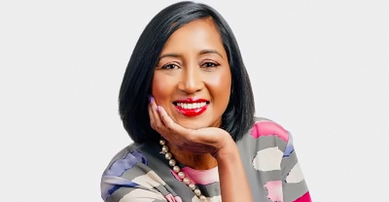October Agencies Smiling woman with shoulder length black hair wearing a colorful striped blouse and pearl necklace resting her chin on her hand while seated