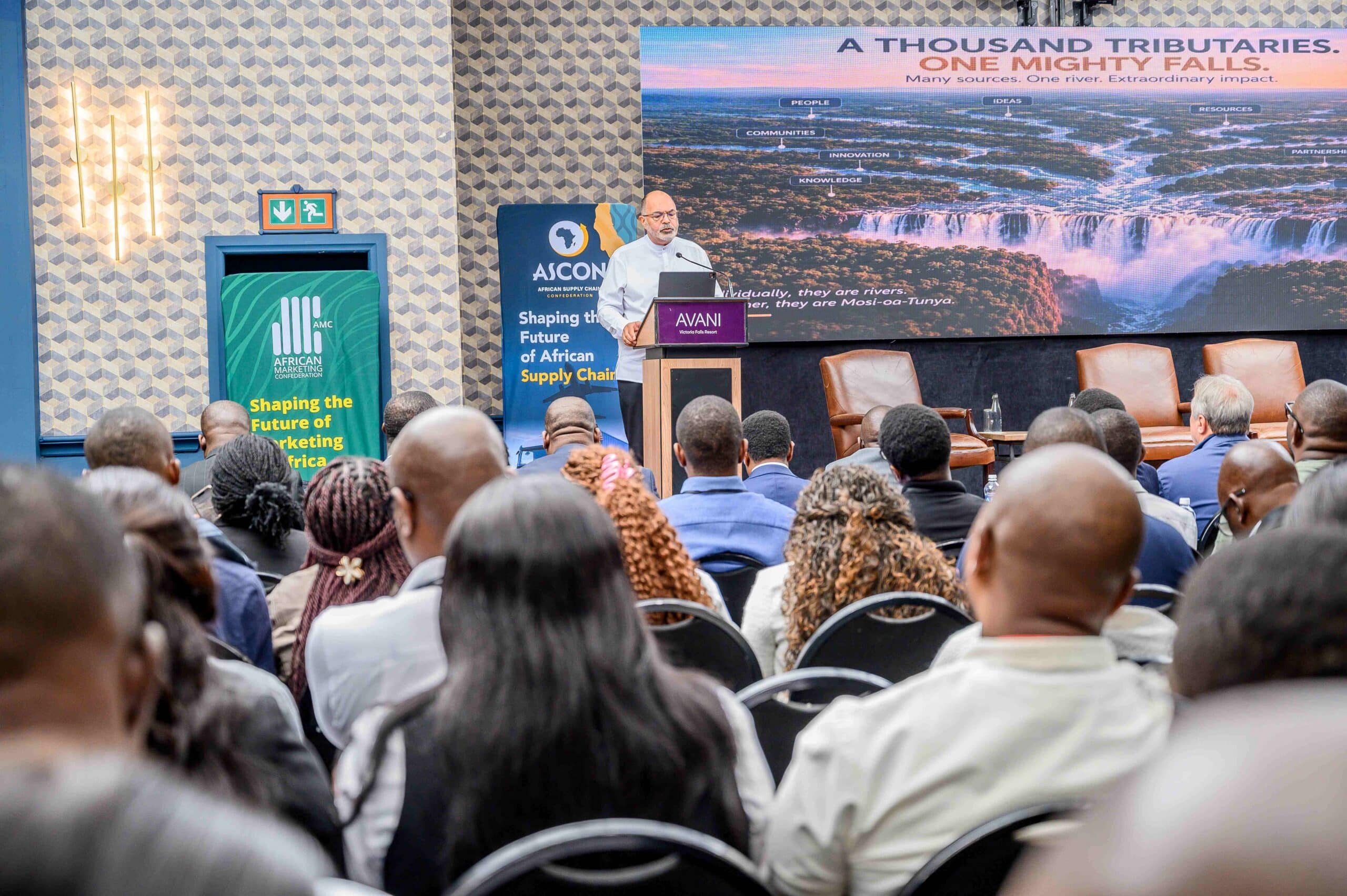 October Conference Speaker at a podium delivering a talk to a seated audience with a large screen showing a waterfall behind and event banners nearby amcascon
