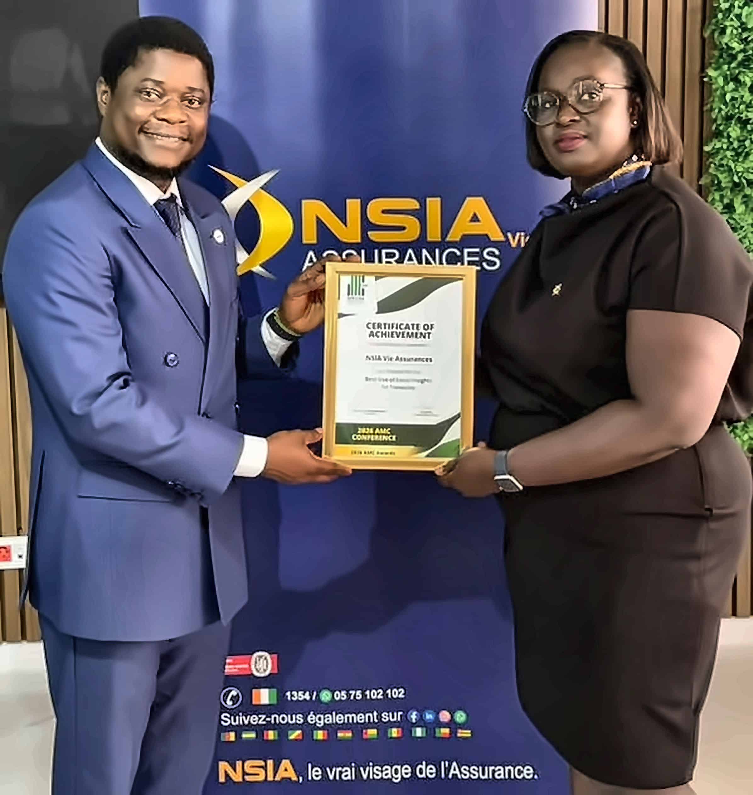 Man in a blue suit hands a framed certificate of achievement to a woman in a black dress both smiling in front of an nsia assurances backdrop
