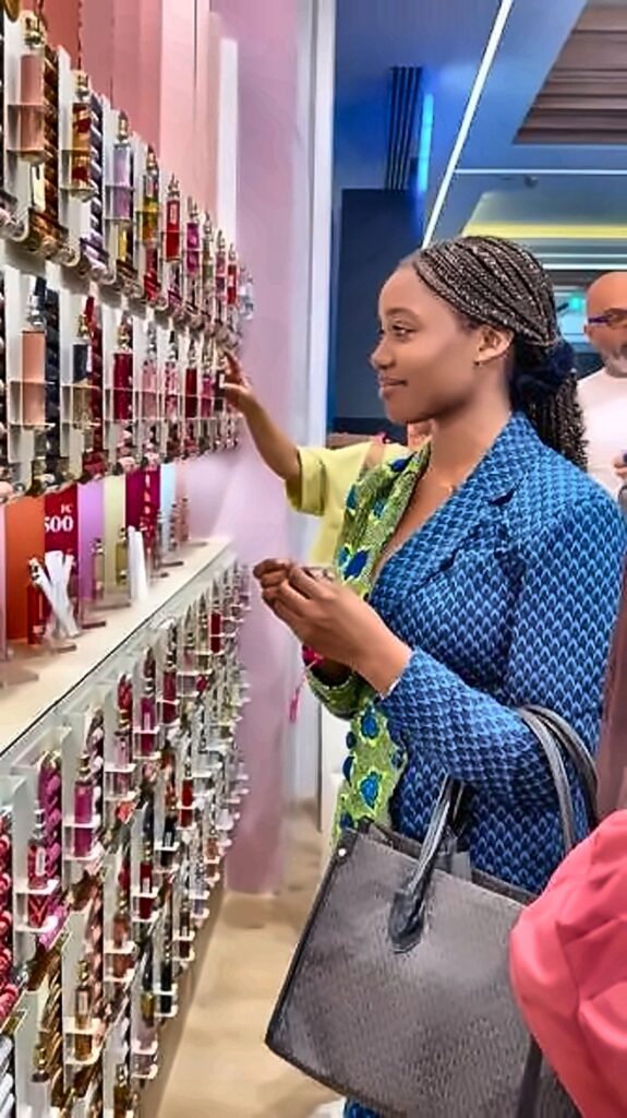 Woman in a blue patterned jacket selecting lipstick from a wall of cosmetics, holding a gray handbag in a store.