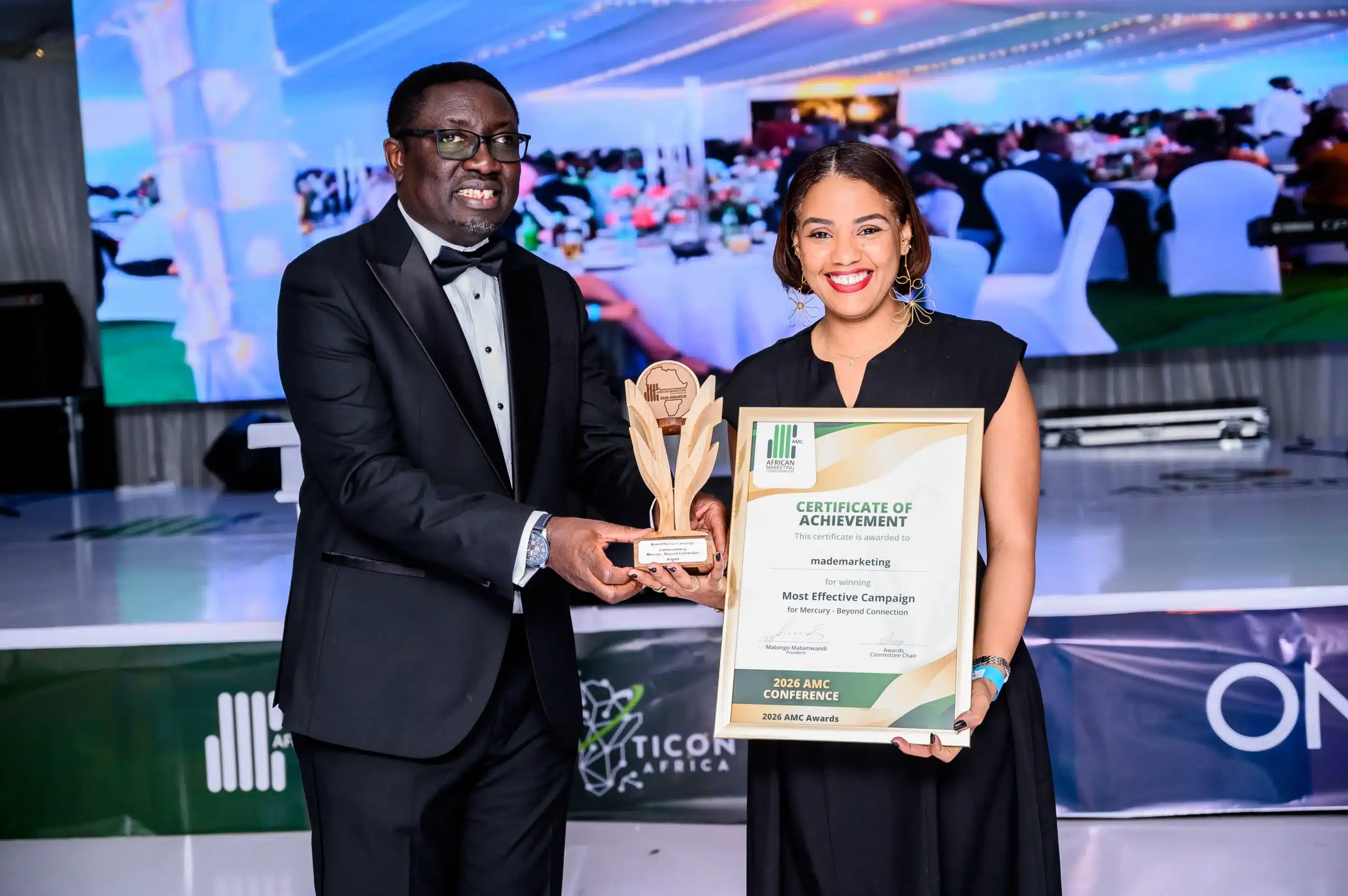 Smiling man in a tuxedo and woman in a black dress pose with a framed certificate and a trophy at an awards ceremony