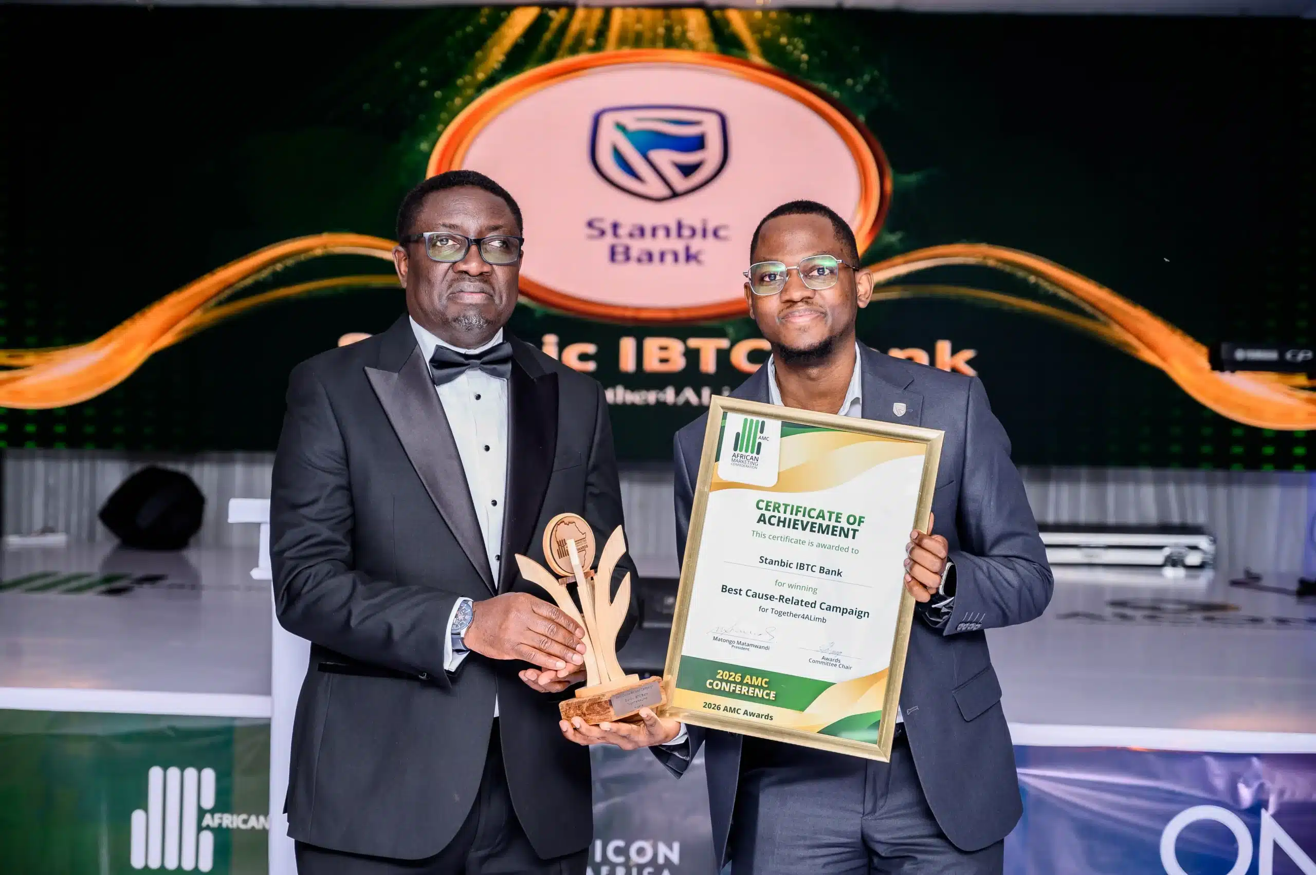 Two men in suits on stage pose with awards one holding a framed certificate the other a wooden trophy with a bank logo backdrop behind them