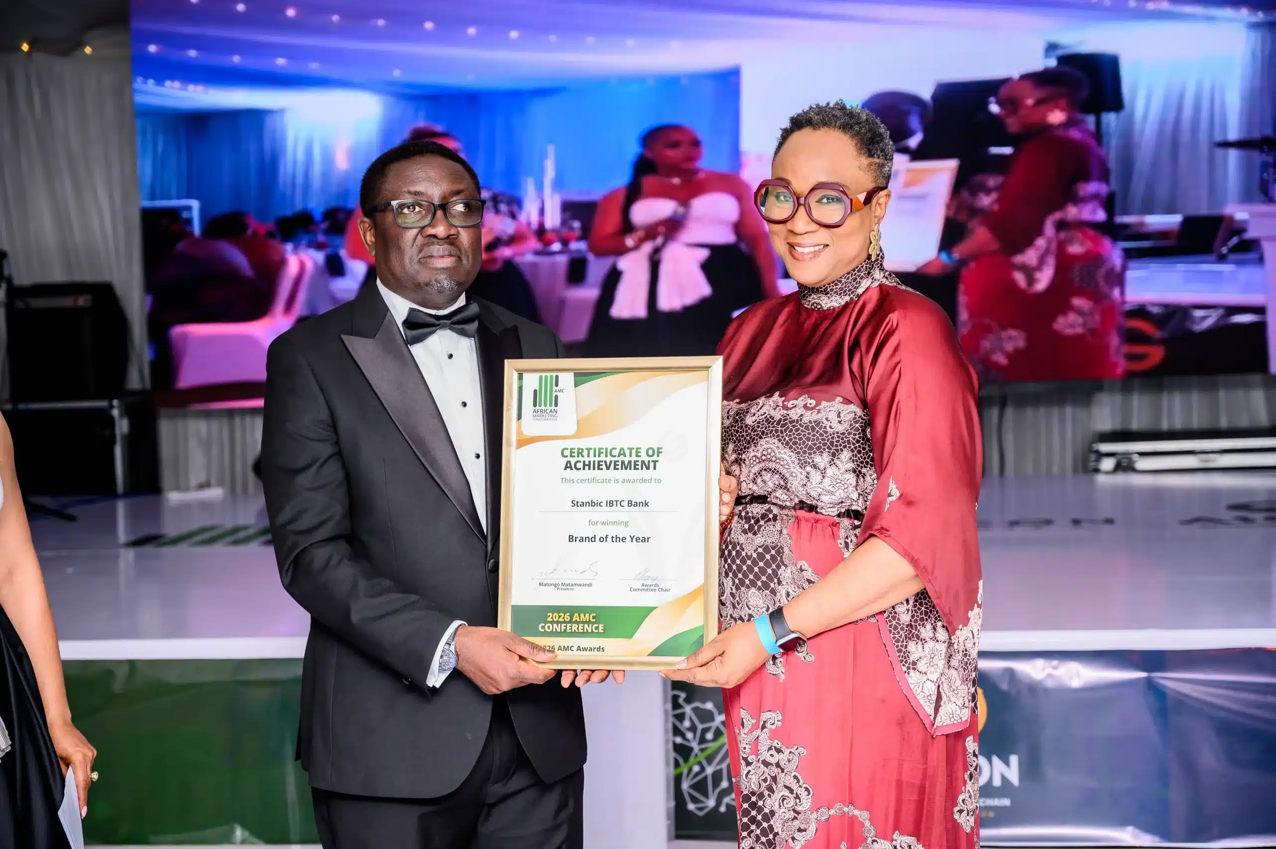 Man in a black tuxedo receives a framed certificate from a woman in a red dress at an awards ceremony