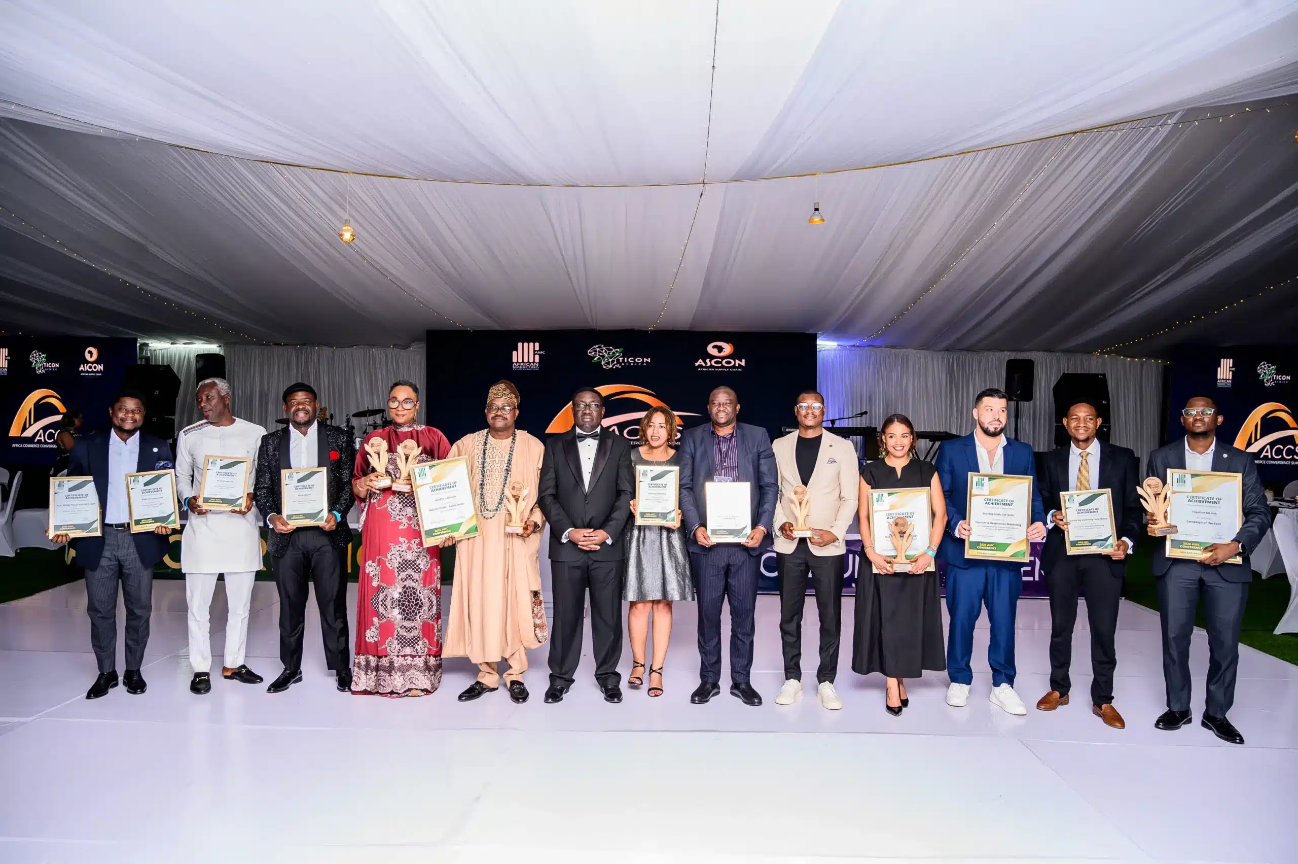 Group of men and women in formal attire posing on a stage each holding a certificate at an awards ceremony under a draped tent
